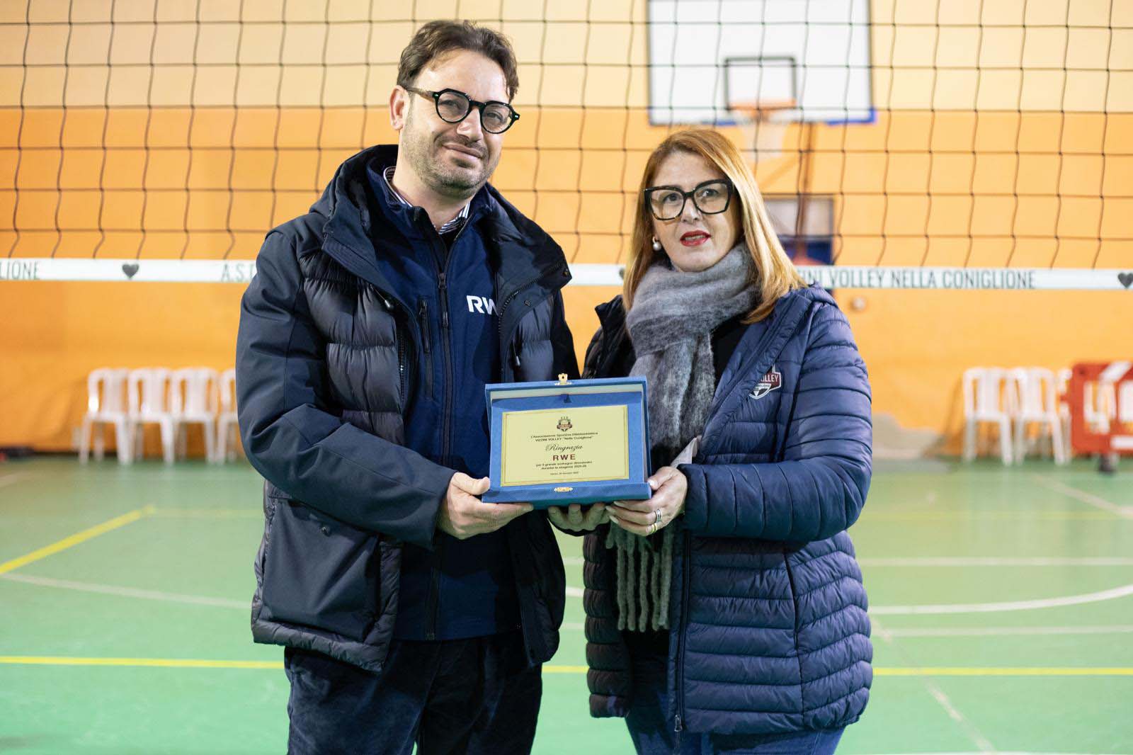 Due persone in palestra, mentre ricevono un premio. Sullo sfondo si vede una rete da pallavolo.