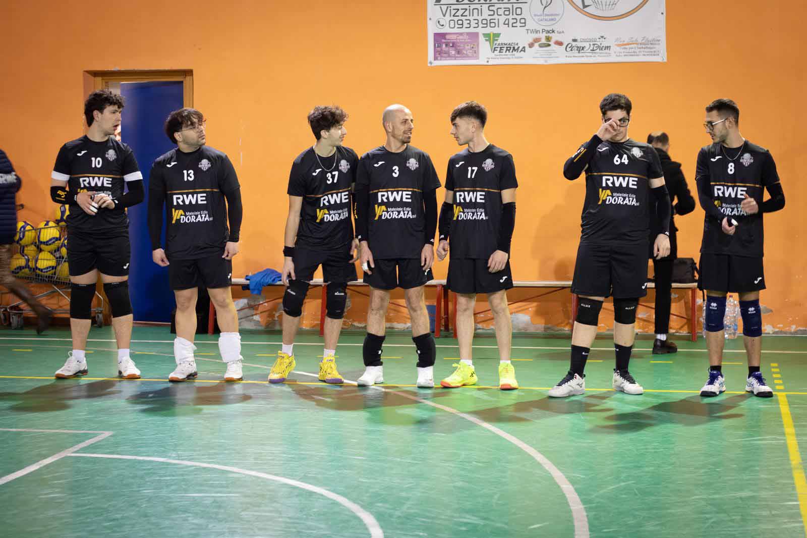 Sei giocatori di pallamano in divisa nera sono schierati su un campo, con sfondo arancione e attrezzature in vista.