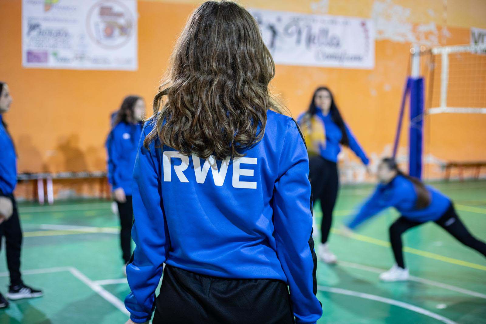 Una ragazza con i capelli mossi indossa una felpa blu con la scritta 'RWE', mentre altre giocano a pallavolo sul campo.