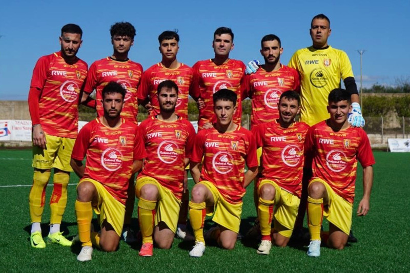 A football team poses in red and yellow kits on a sunny day on the pitch, showcasing team spirit and camaraderie.