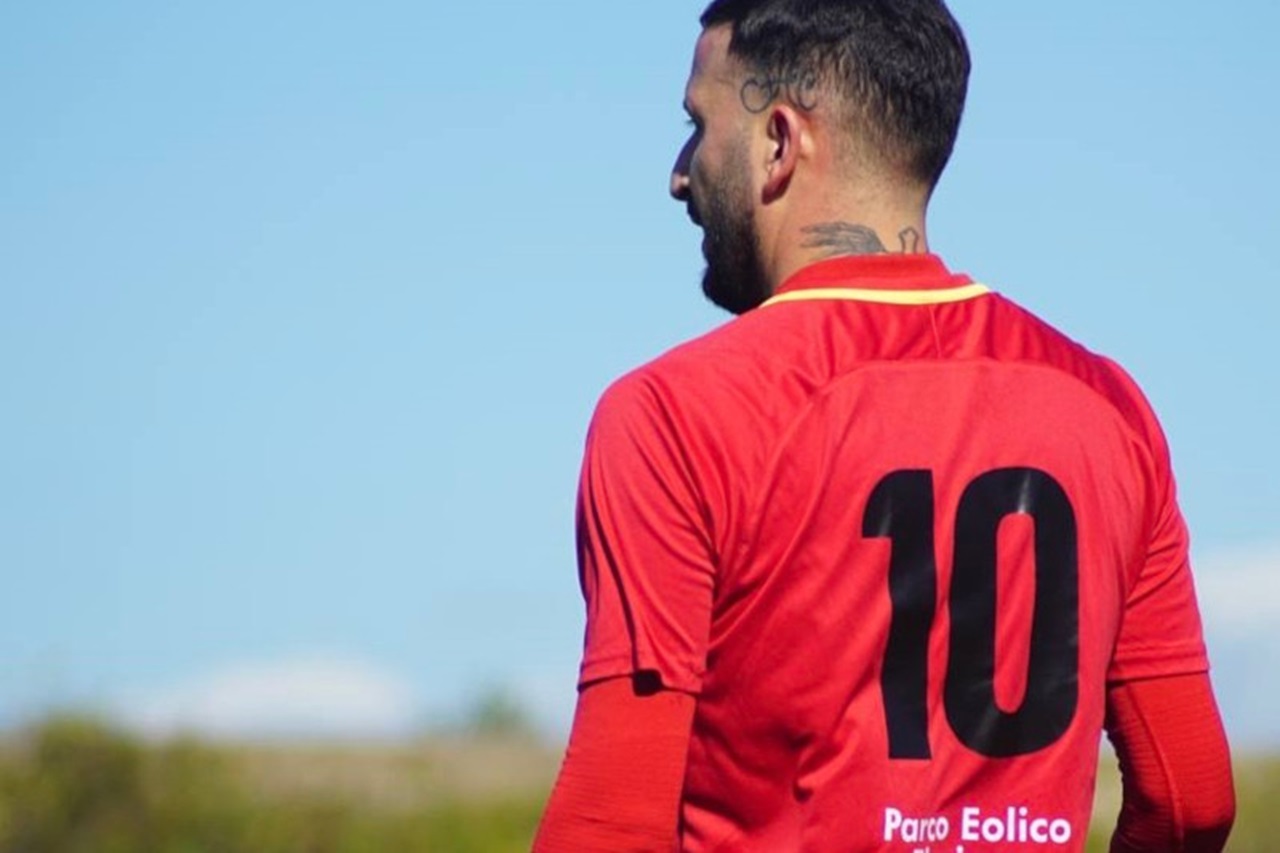 Un calciatore in maglia rossa, numero 10, sta in piedi di spalle di fronte a un cielo azzurro e limpido.