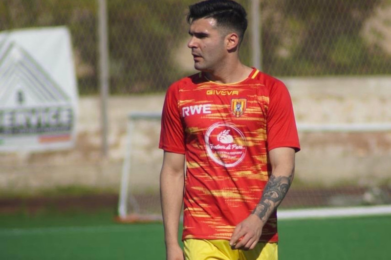 A football player stands on the pitch, wearing a red and yellow team jersey with a tattoo on his arm.