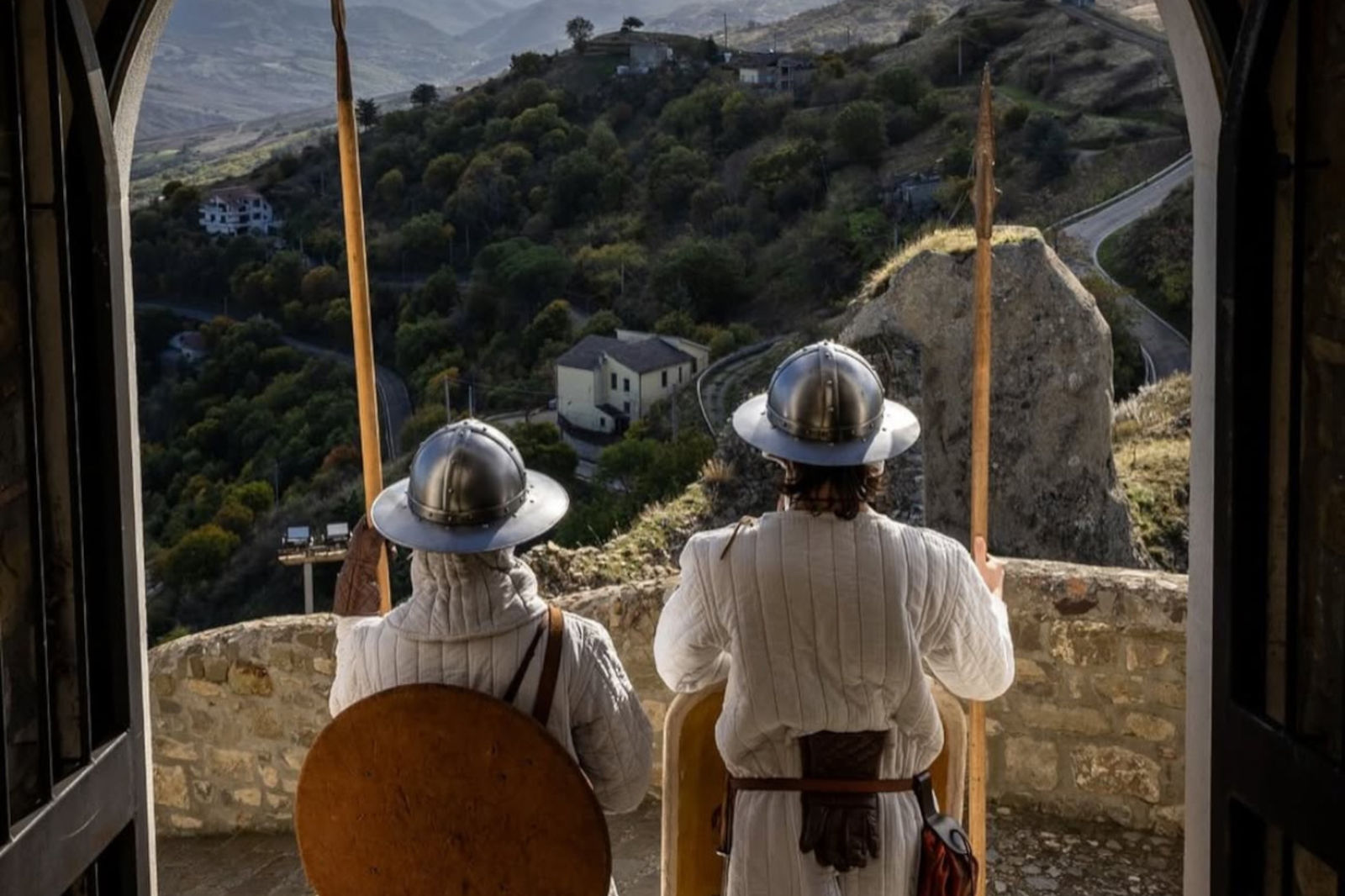 Two armoured guards stand at a stone balcony, gazing over a hilly landscape dotted with houses and winding roads.
