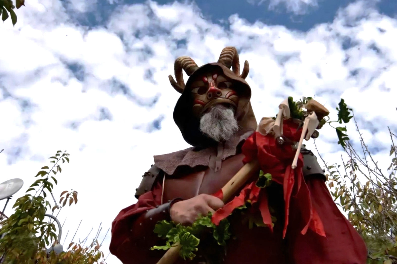 A masked person in a red cloak holds a staff adorned with greenery and a puppet, surrounded by a cloudy sky.