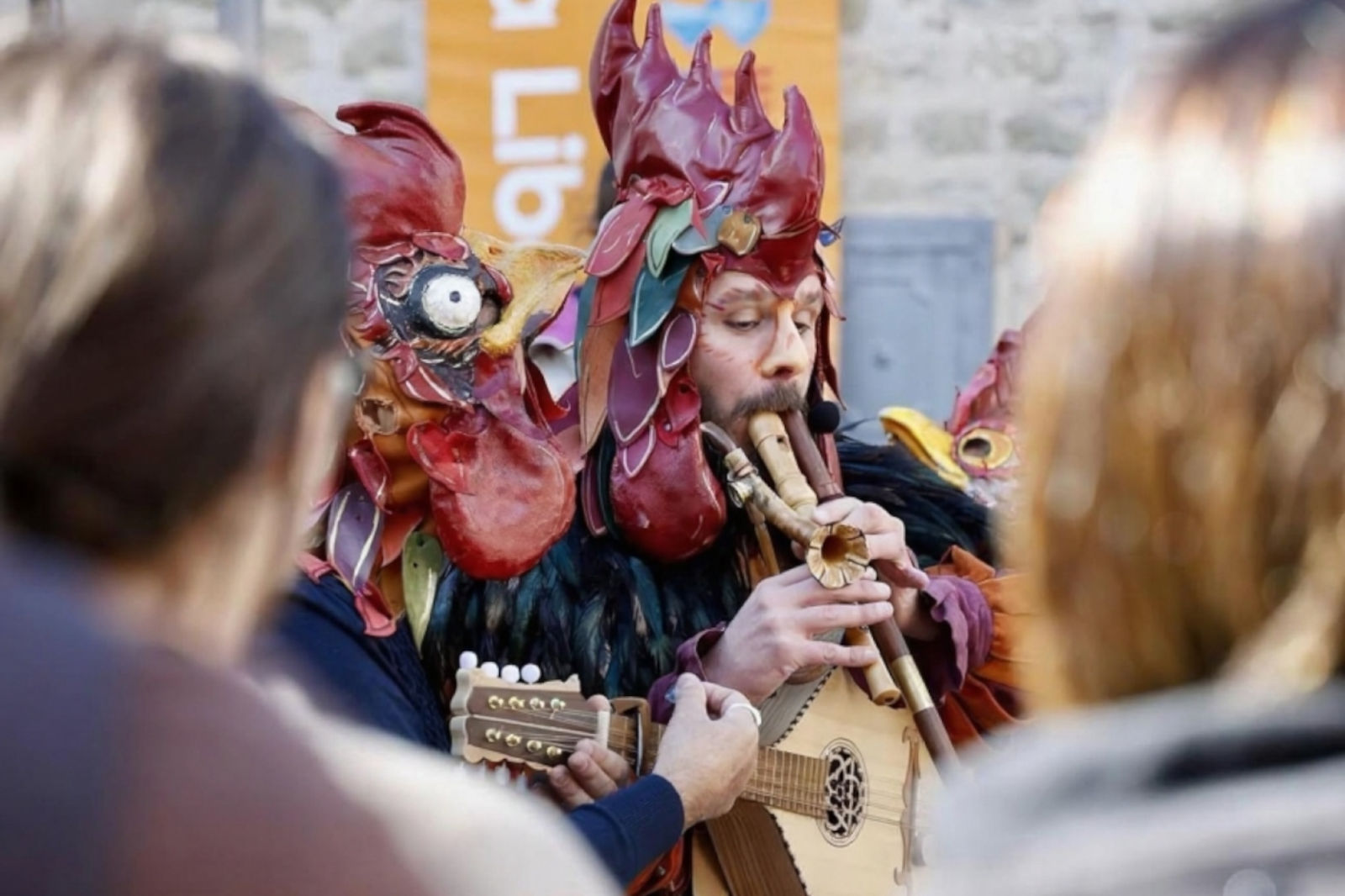 Un artista con una maschera di gallo colorata suona un strumento a corda mentre altri godono dell'atmosfera vivace.