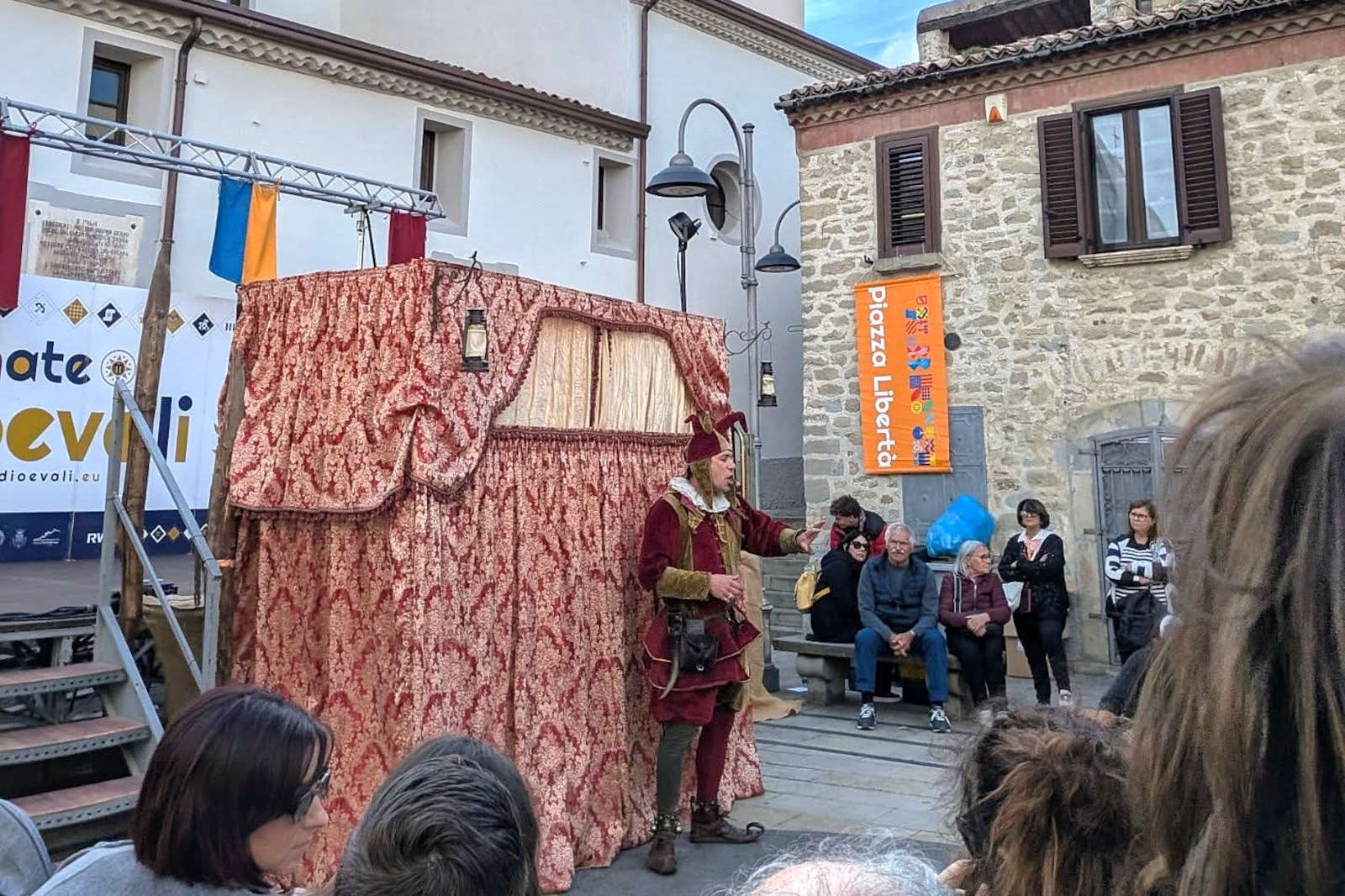 Un artista in costume rosso gesticola di fronte a un drappo scenico ornato, con un pubblico seduto nelle vicinanze.