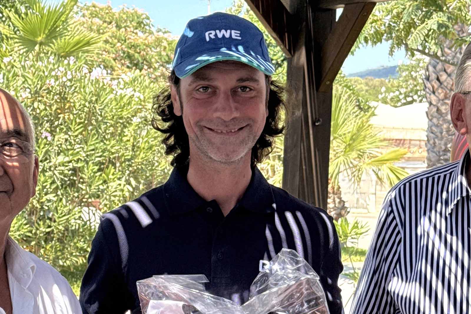 Uomo sorridente con cappellino navy con la scritta «RWE» e polo tiene un pacco in cellophane sotto una pergola di legno tra palme.