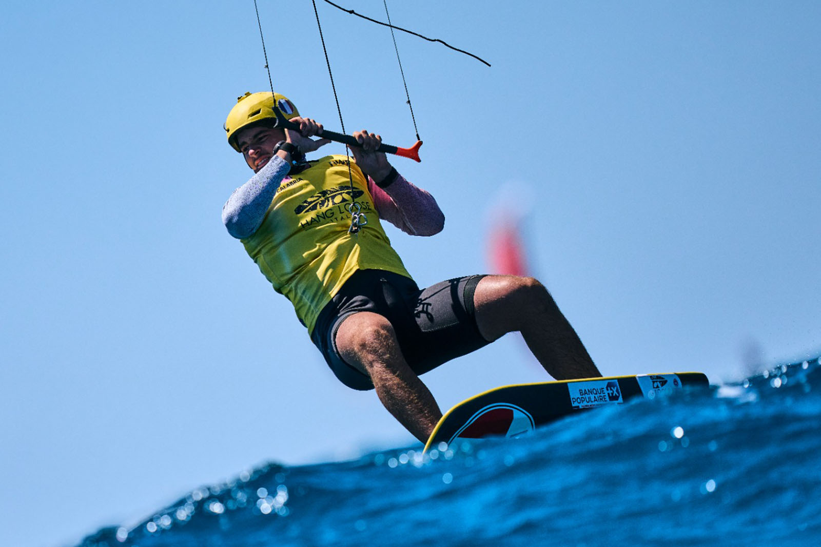 Un kitesurfer con casco e pettorina gialli si inclina su una tavola tenendo una barra di controllo rossa sopra il mare mosso e cielo sereno.