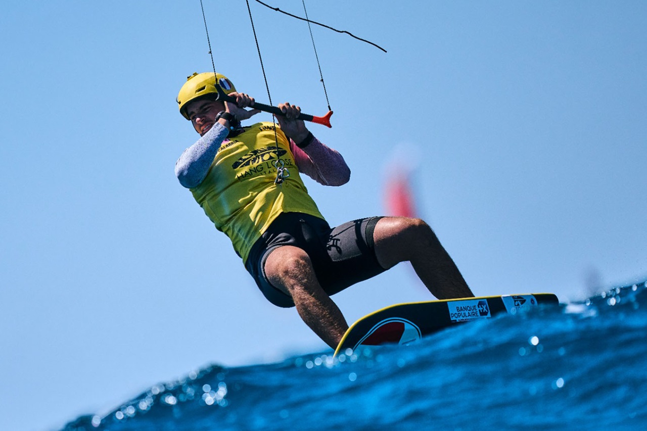 Un kitesurfer con casco e pettorina gialli si inclina su una tavola tenendo una barra di controllo rossa sopra il mare mosso e cielo sereno.