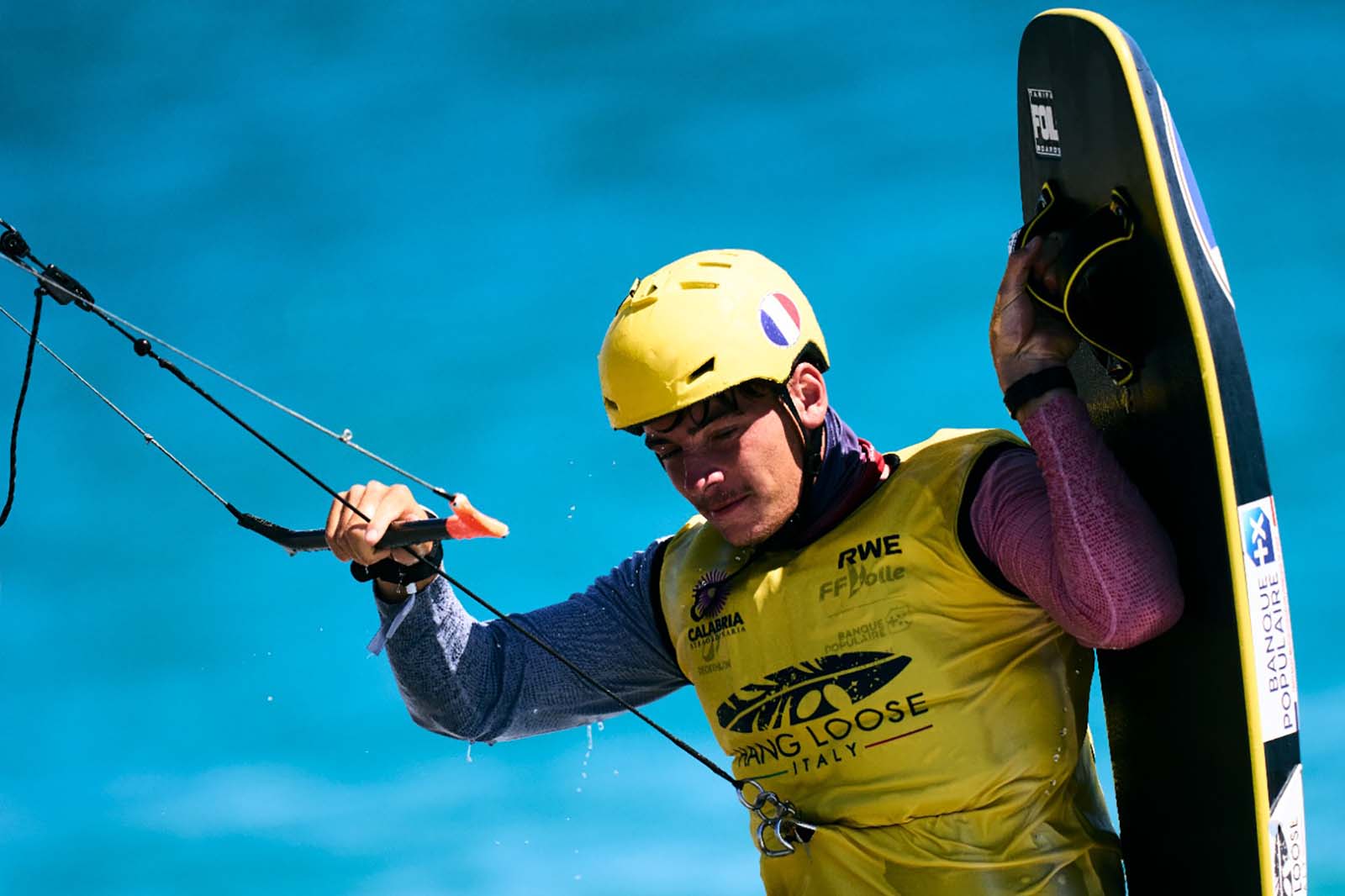 Un uomo con casco giallo e giubbotto giallo etichettato «LONG LOOSE» tiene una tavola da wake nera e una maniglia di traino sopra acqua turchese.