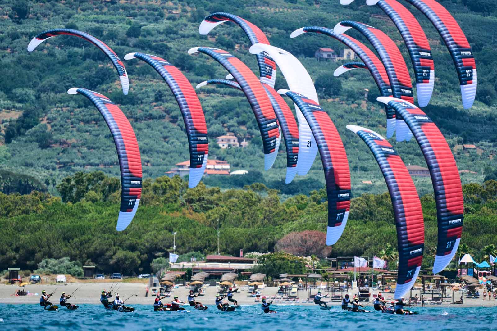 Aquiloni gonfiabili rossi, neri e bianchi curvano sopra acqua azzurra mentre numerosi kiteboarder su hydrofoil sfrecciano vicino a una spiaggia sabbiosa e colline verdi.