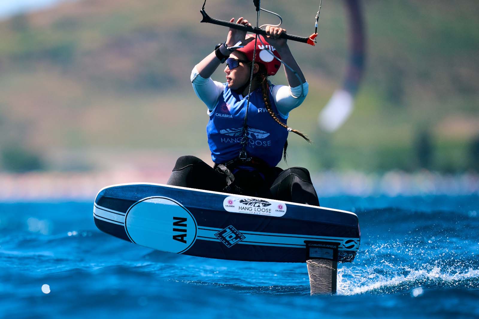 Una kitesurfer con casco rosso e giubbotto blu plana sopra acque blu intense su una tavola foil azzurra con la scritta «AIN» e «HANG LOOSE».