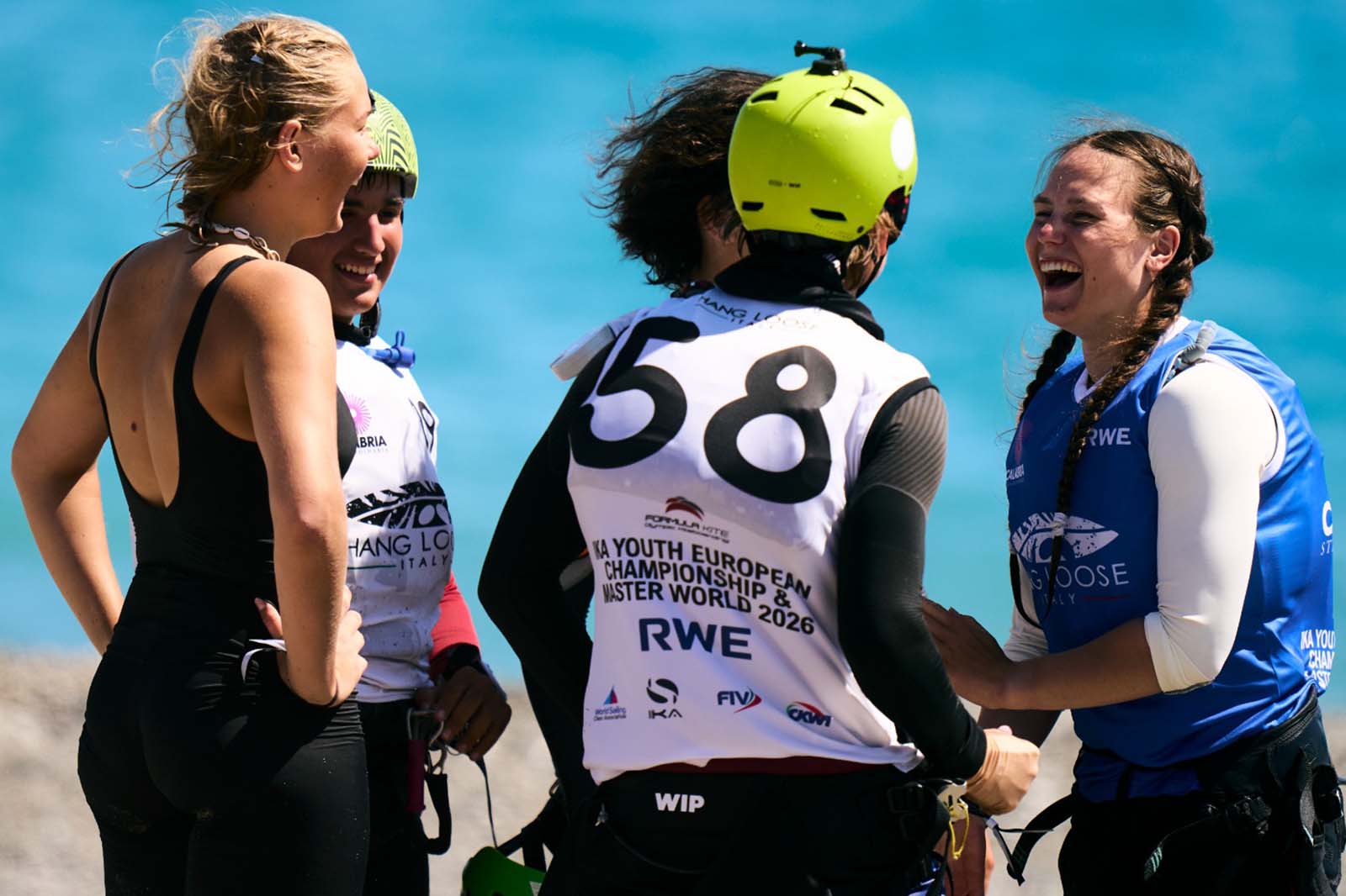 Quattro velisti in mute e giubbotti salvagente stanno su una spiaggia sabbiosa davanti a acqua turchese, sorridendo; un giubbotto reca "58" e un altro "RWE".