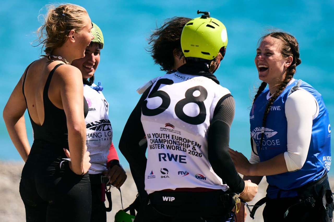 Quattro velisti in mute e giubbotti salvagente stanno su una spiaggia sabbiosa davanti a acqua turchese, sorridendo; un giubbotto reca "58" e un altro "RWE".