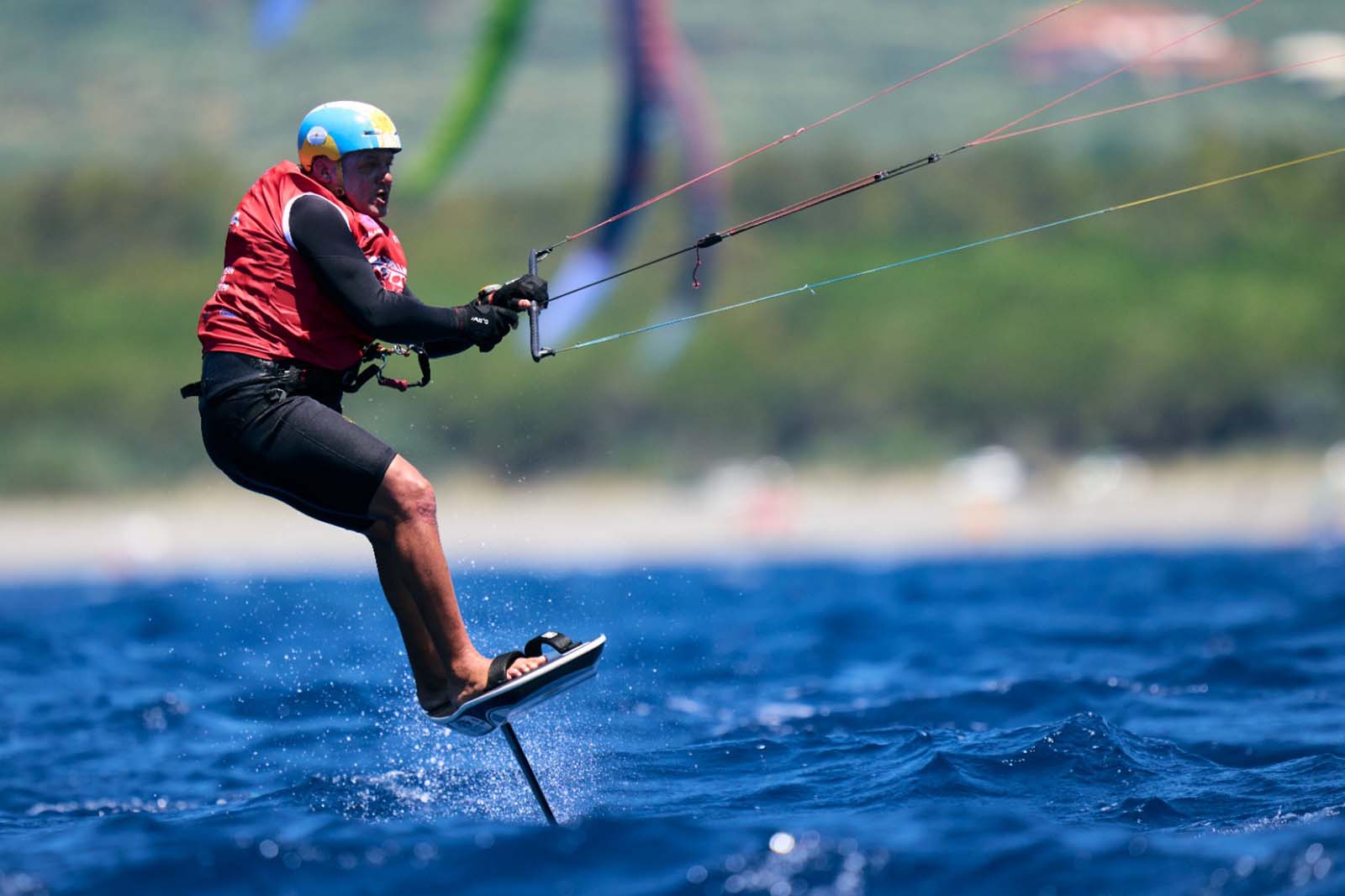 Un uomo con giubbotto rosso e casco blu sorvola l'acqua mossa su una tavola idrofoil, tenendo le linee colorate del kite.