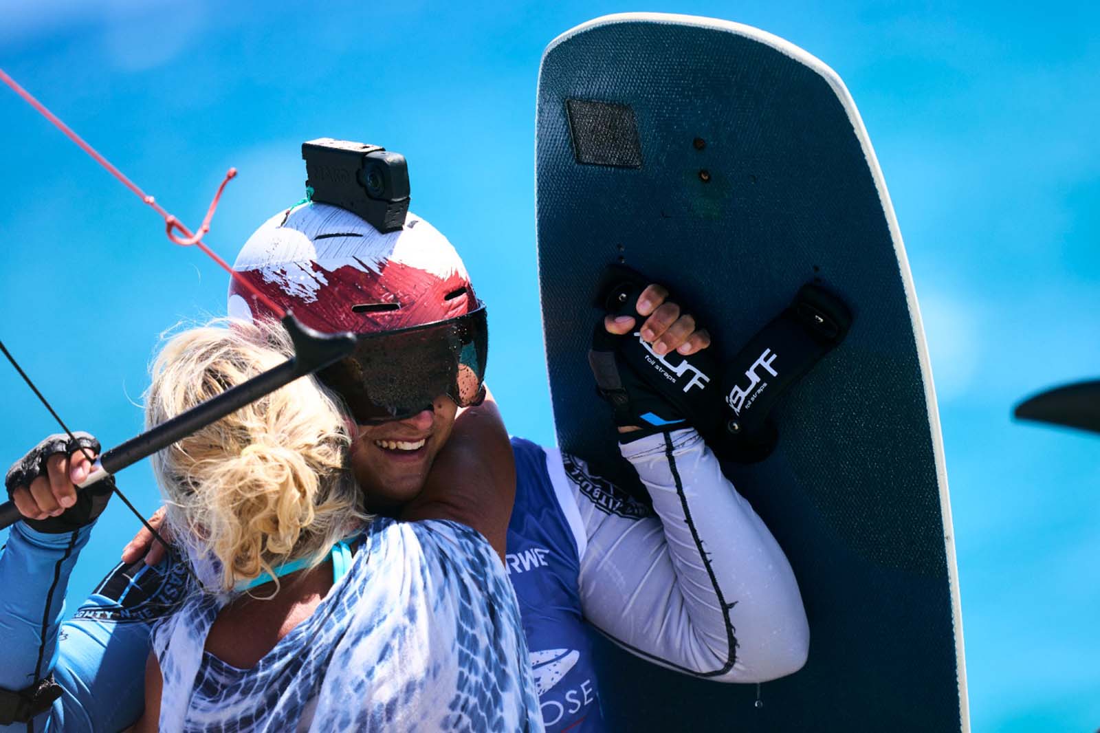 Un atleta acquatico sorridente con casco rosso e maniche in neoprene bianche tiene una tavola nera mentre abbraccia una donna bionda davanti ad acqua turchese.