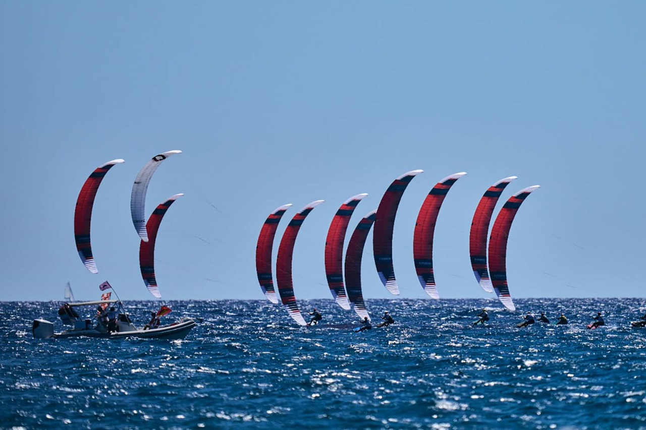 Più aquiloni curvi rossi e bianchi solcano un mare blu mosso, con kitesurfer in silhouette e una piccola barca a motore con bandiera.