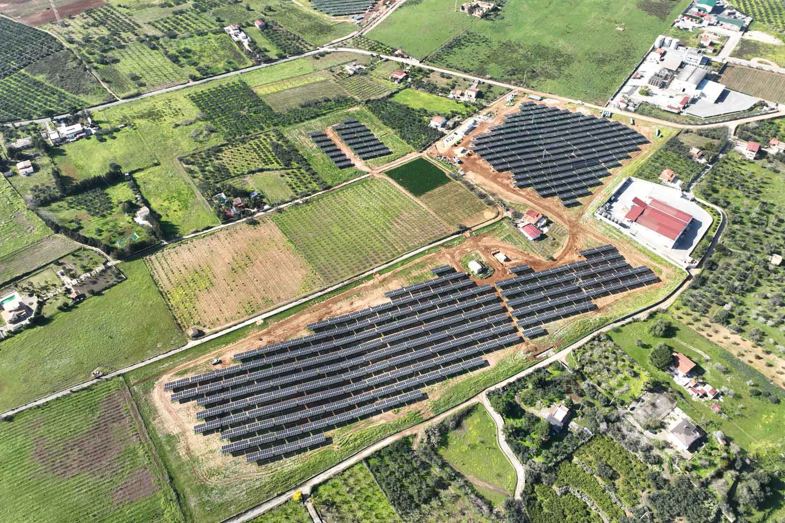 An aerial view of solar panels arranged in rows on farmland, surrounded by green fields and small settlements.
