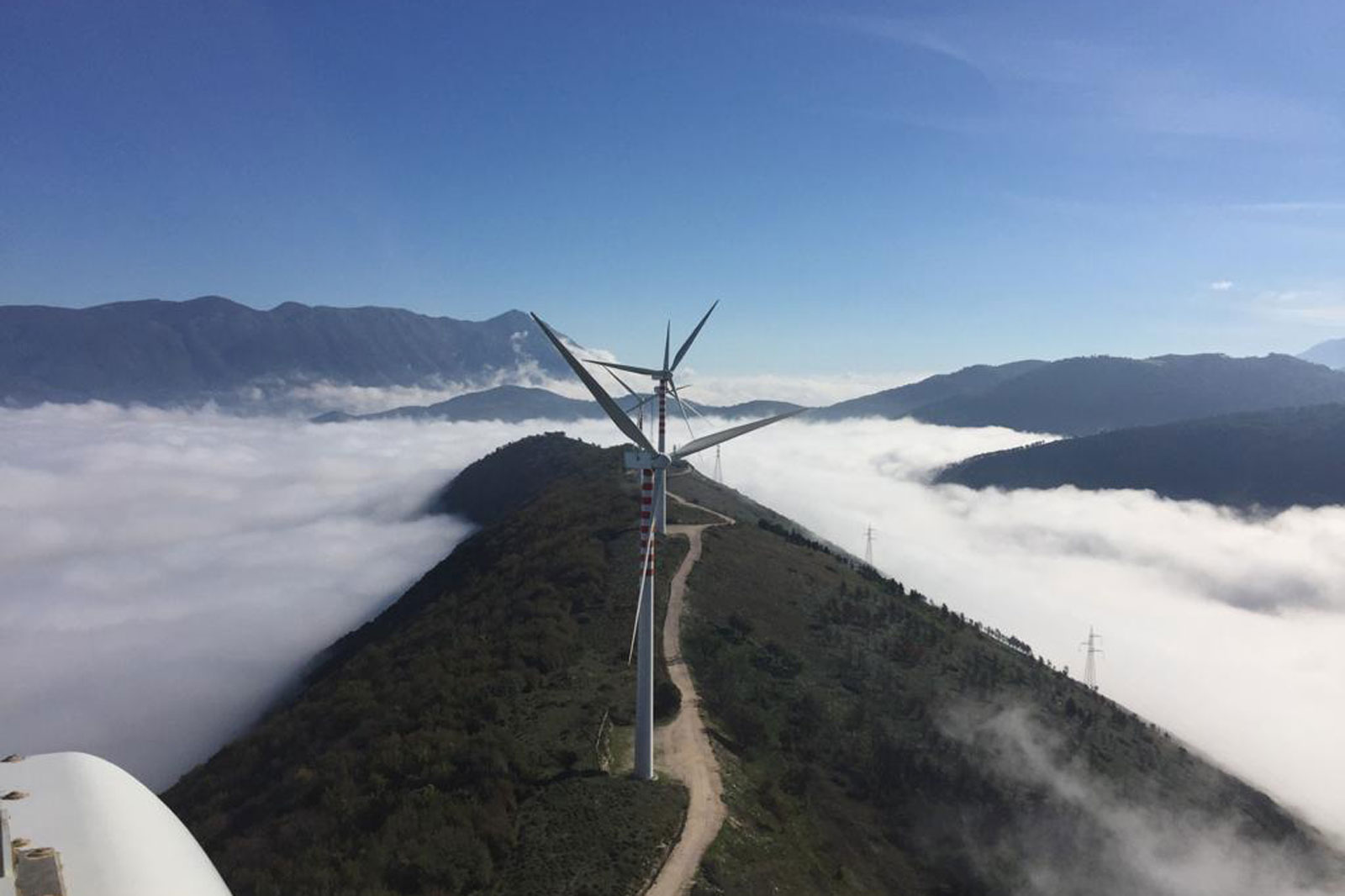 A panoramic view of multiple wind turbines on a mountain peak, surrounded by clouds with hills and power lines in the background.