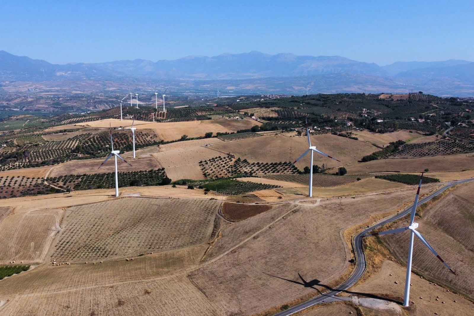Aerial view of rolling hills with wind turbines, olive groves, and a winding road under a clear blue sky.
