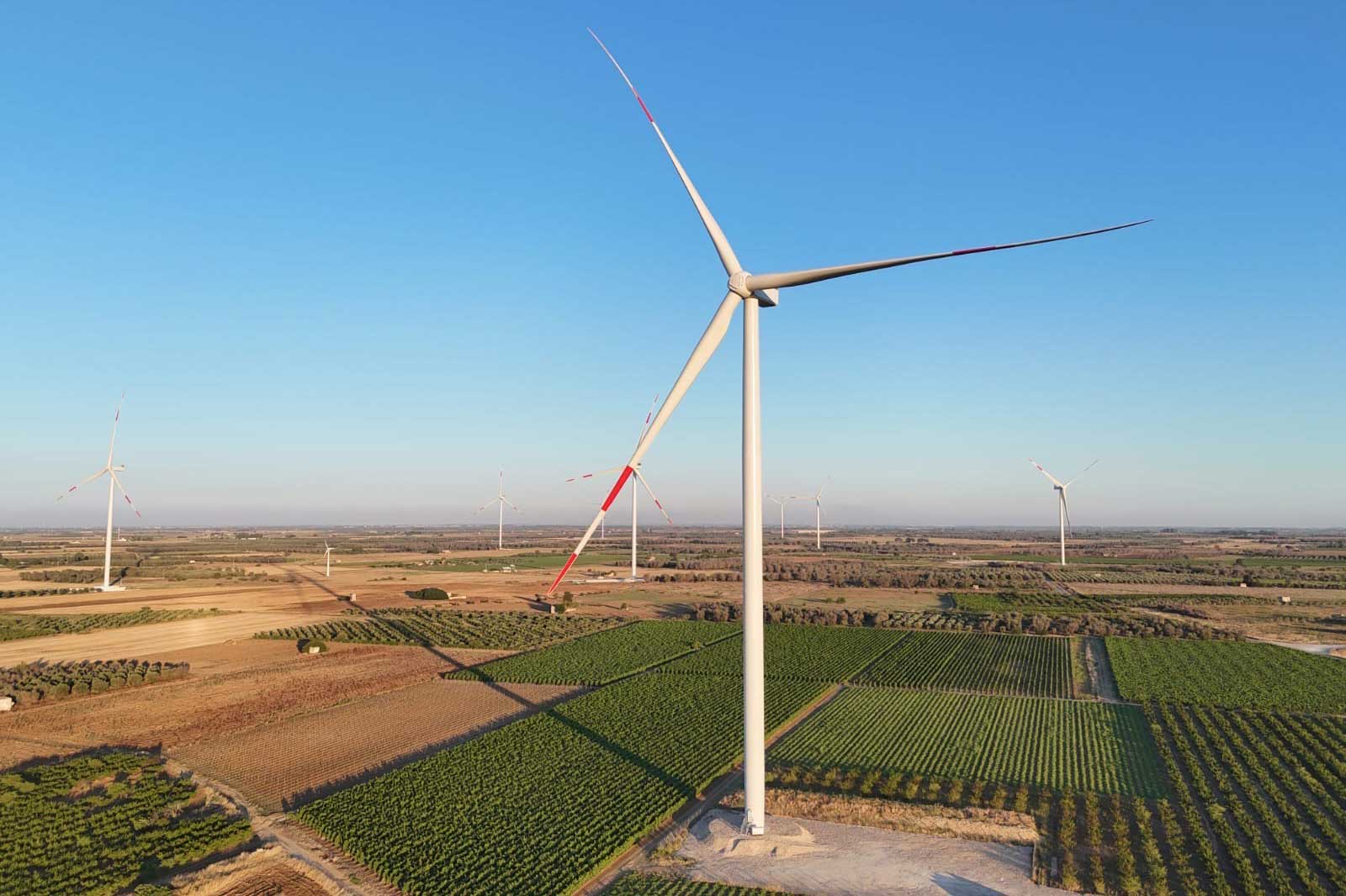 A wind turbine stands in a vast field with green vineyards and golden crops beneath a clear blue sky.