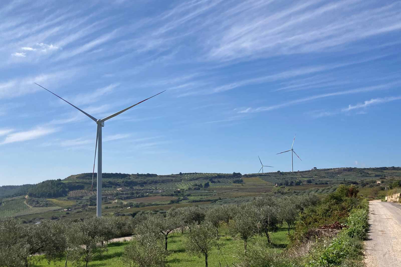A scenic view featuring three wind turbines on a hillside, surrounded by olive trees and fields under a clear blue sky.