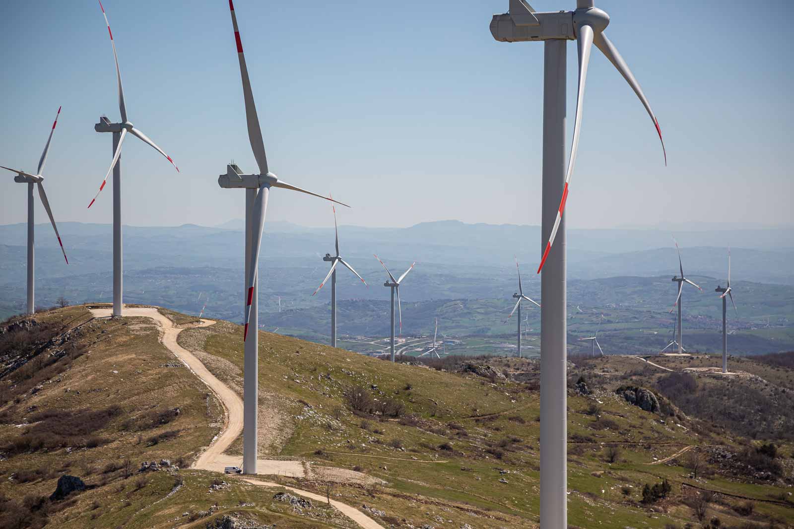 A panoramic view of wind turbines on a hill, with a dirt path leading through green grass and scattered rocks under a clear blue sky.
