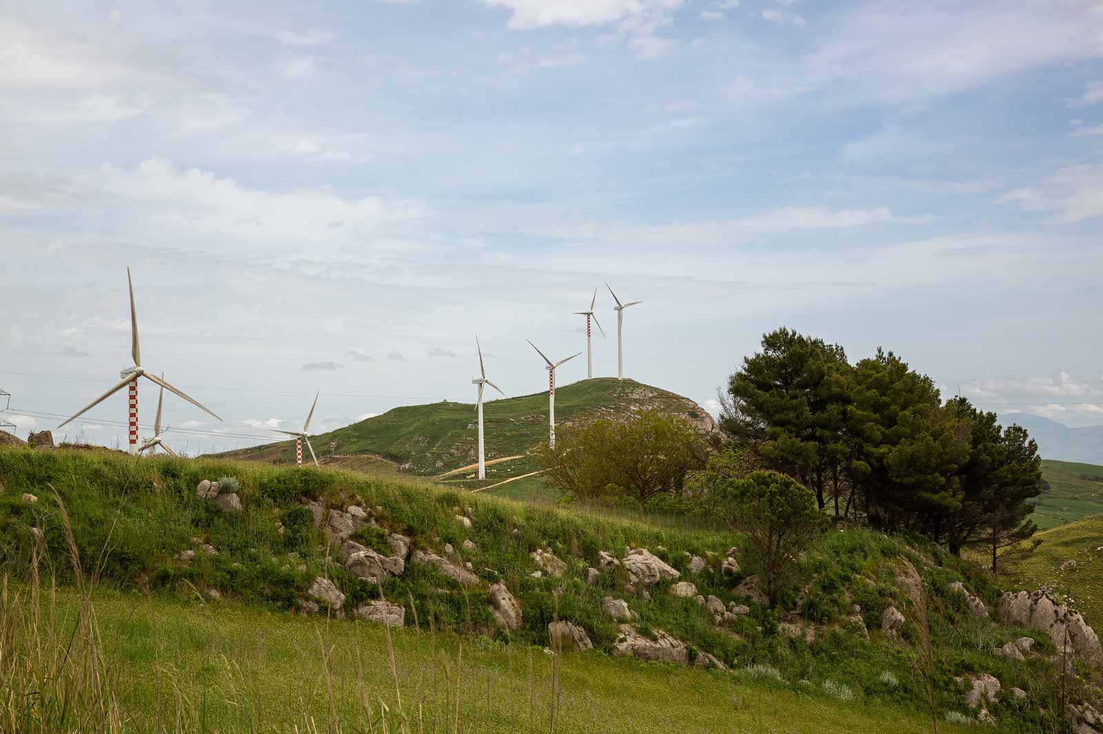 A scenic view of wind turbines on a hillside, surrounded by greenery and rocks under a partly cloudy sky.