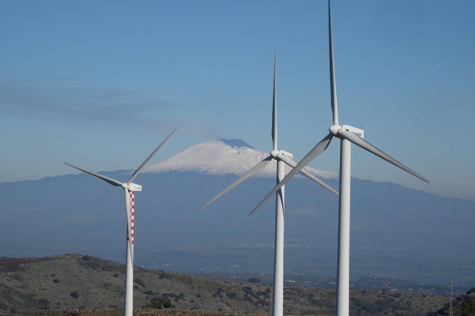 Three wind turbines stand in the foreground, with Mount Etna covered in snow in the background against a clear blue sky.