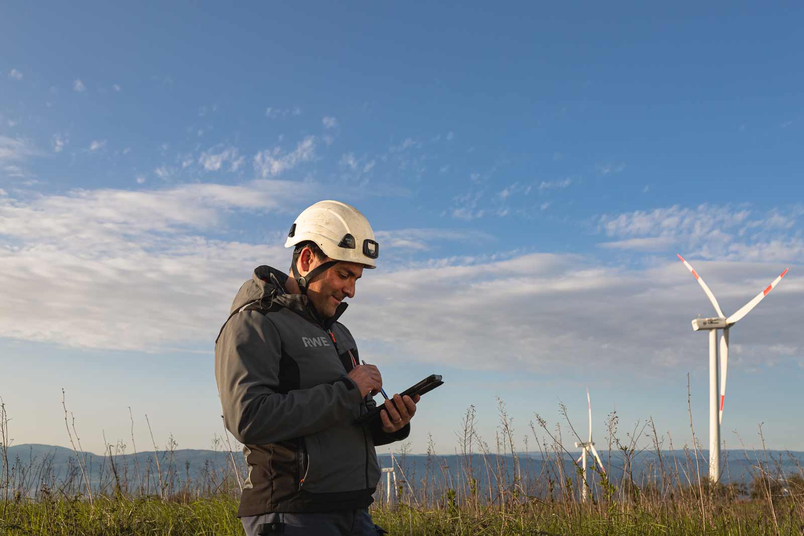 Un lavoratore con un elmetto di sicurezza usa un tablet all'aperto, con turbine eoliche e un cielo blu nel background.