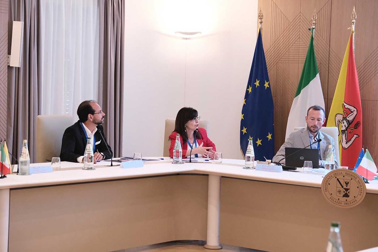 Un incontro con funzionari a un tavolo rotondo, con bandiere di Italia, UE e Sicilia, laptop e bottiglie d'acqua presenti.