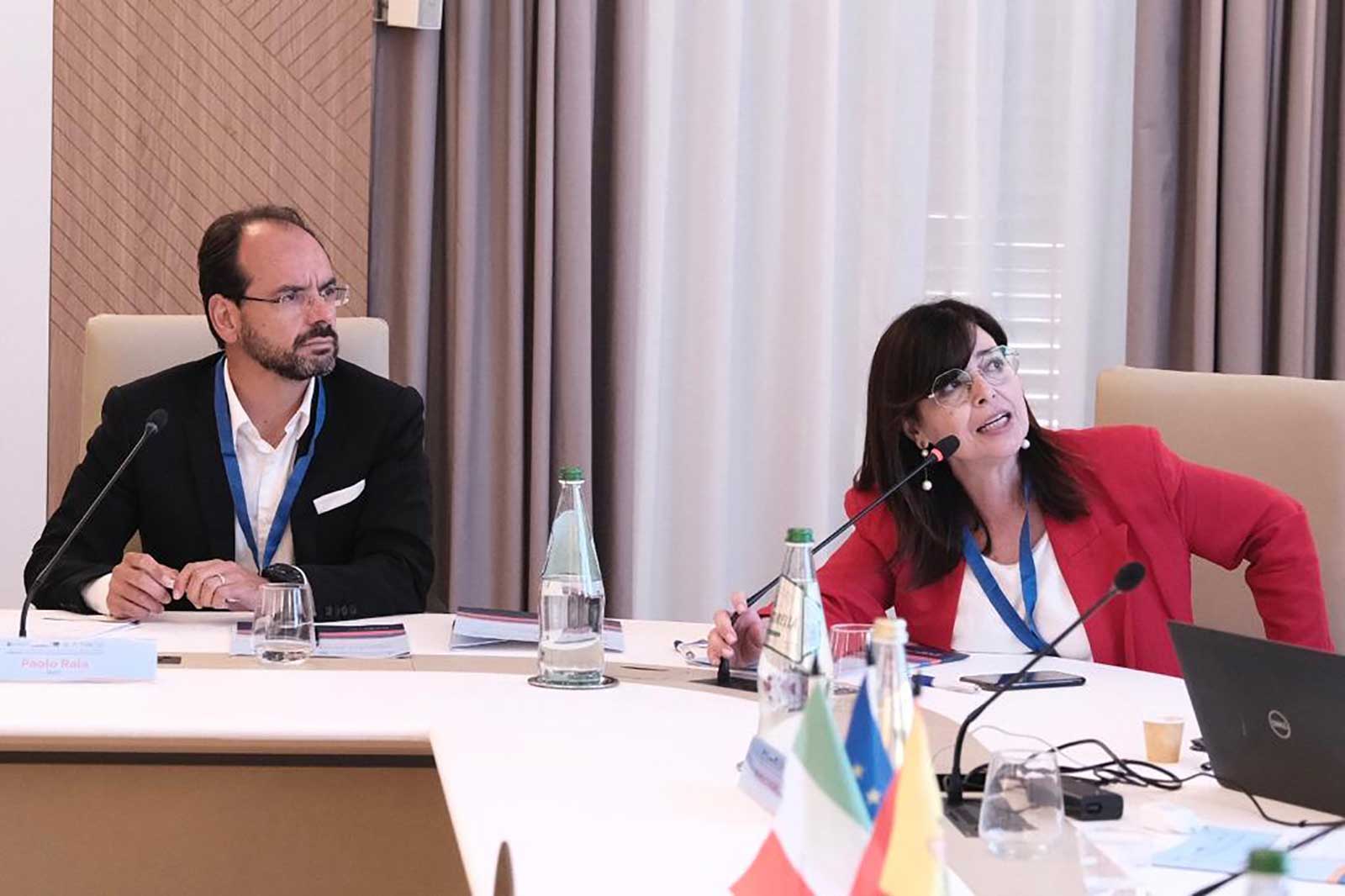Due professionisti impegnati in una discussione a un tavolo di conferenza con bottiglie d'acqua e bandiere in vista.