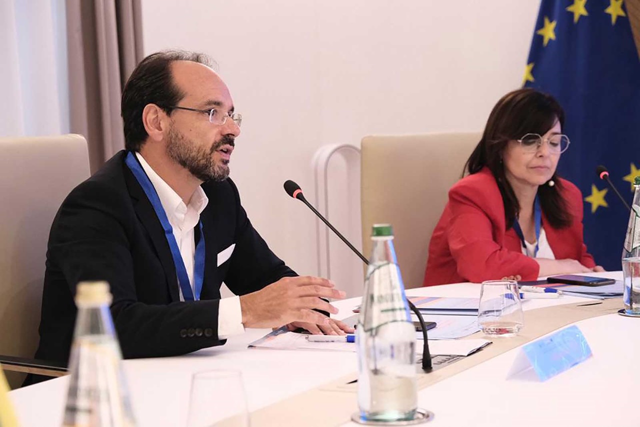 Due persone sedute a un tavolo di conferenza, impegnate in una discussione con documenti e una bottiglia d'acqua presenti.
