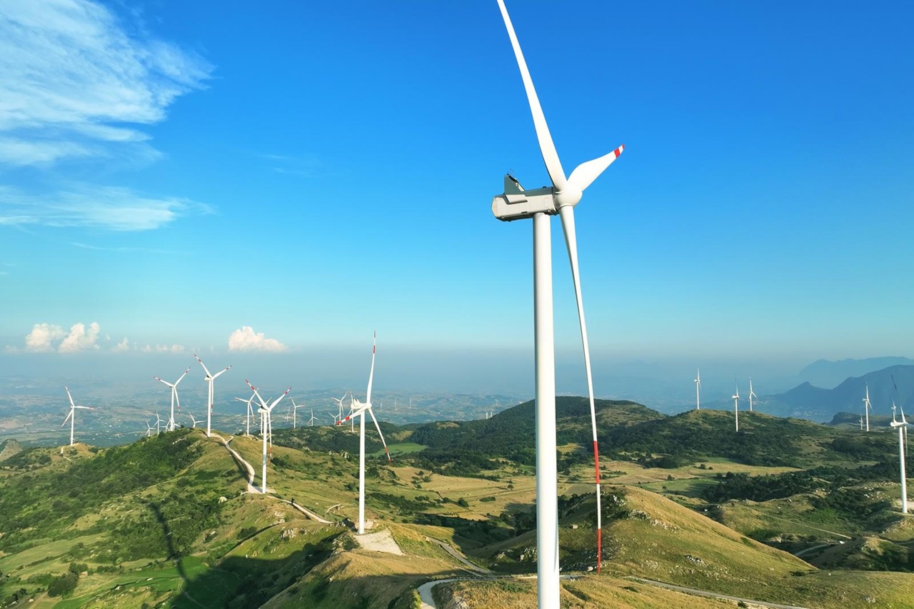 A panoramic view of wind turbines on rolling green hills under a clear blue sky.