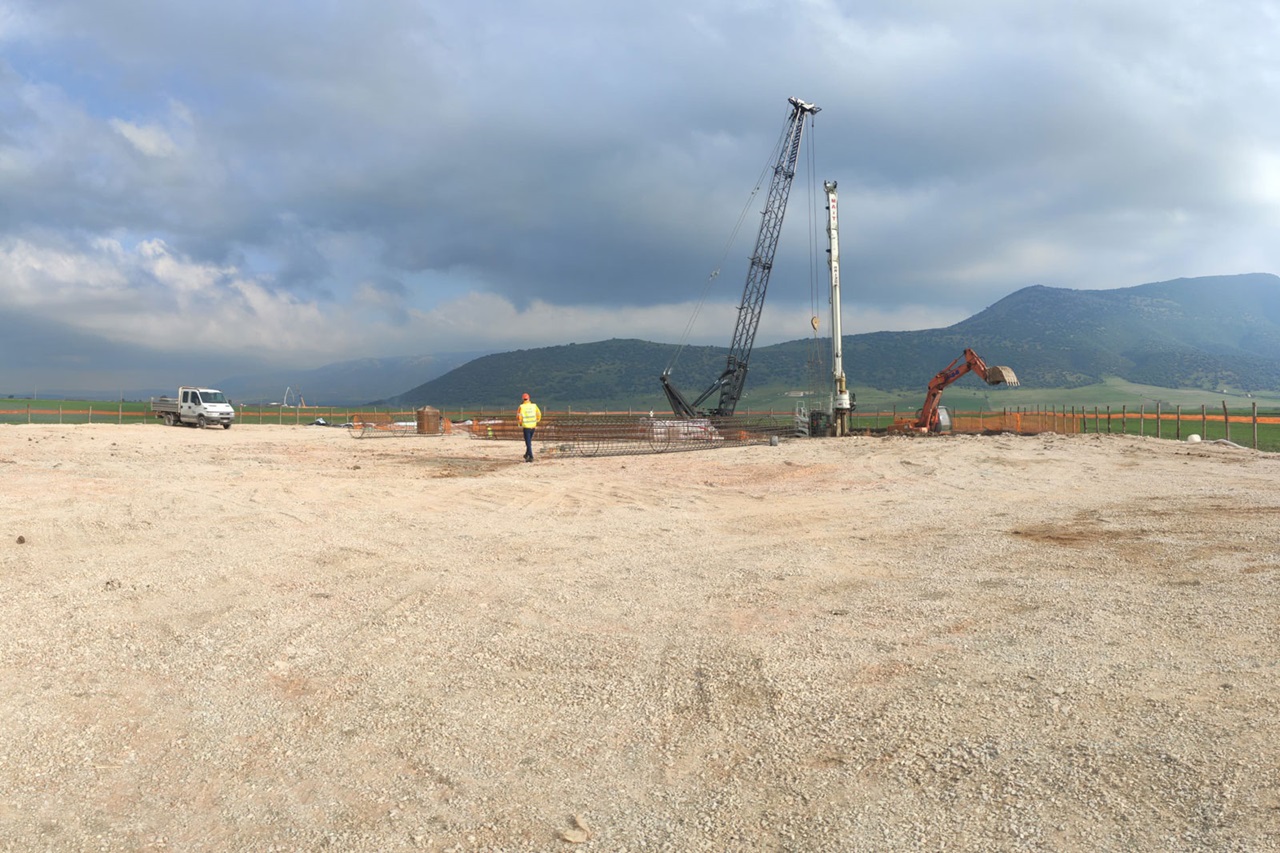 Un cantiere con macchinari, terreno di ghiaia e un lavoratore in giubbotto di sicurezza. Montagne e cieli nuvolosi sullo sfondo.