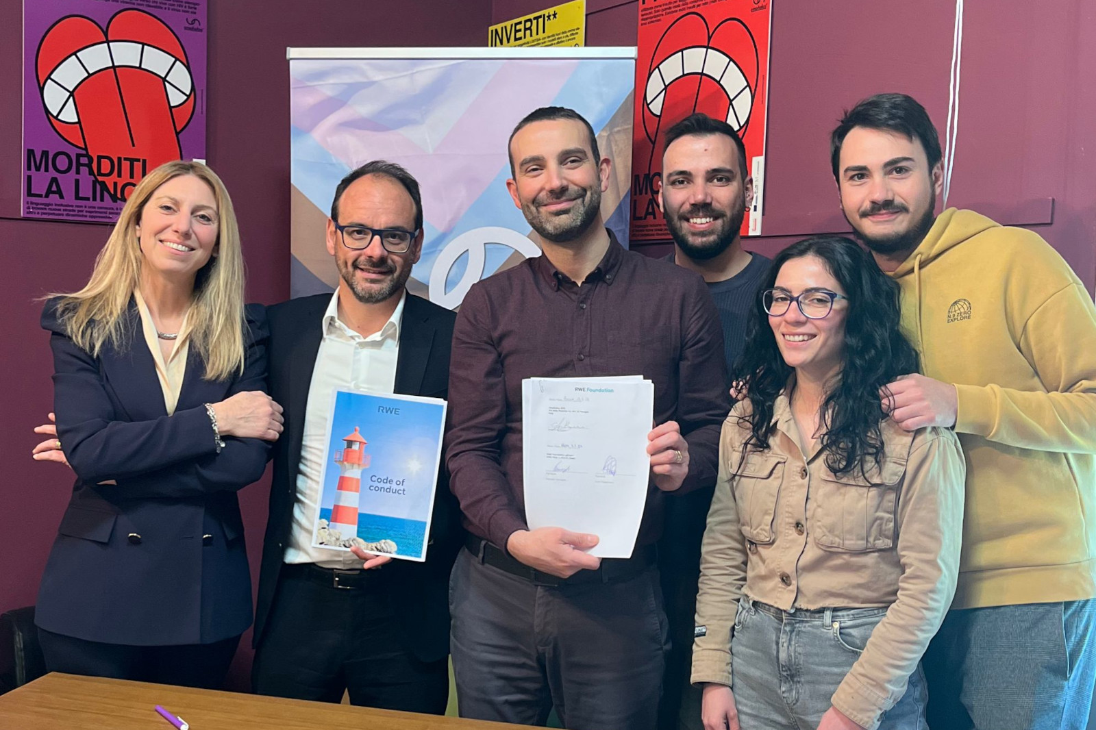 A group of individuals standing together, holding documents, with colourful posters in the background.