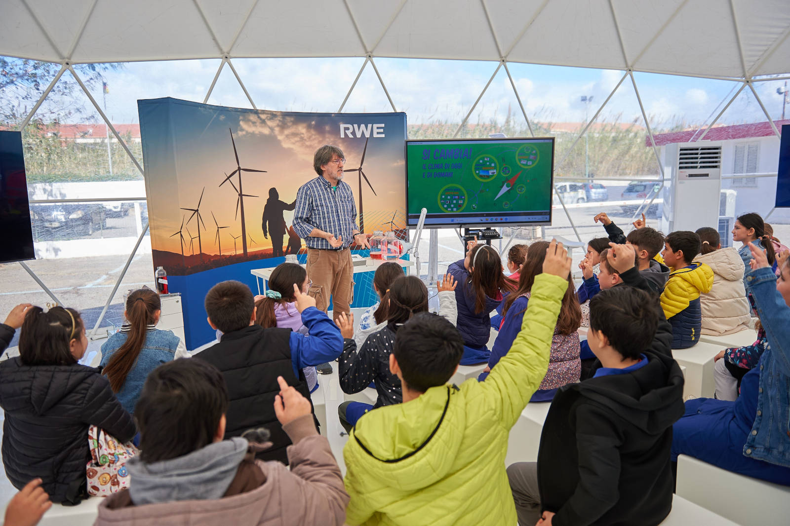 Un gruppo di bambini coinvolti in una presentazione interattiva sulle energie rinnovabili, con turbine eoliche sullo sfondo.