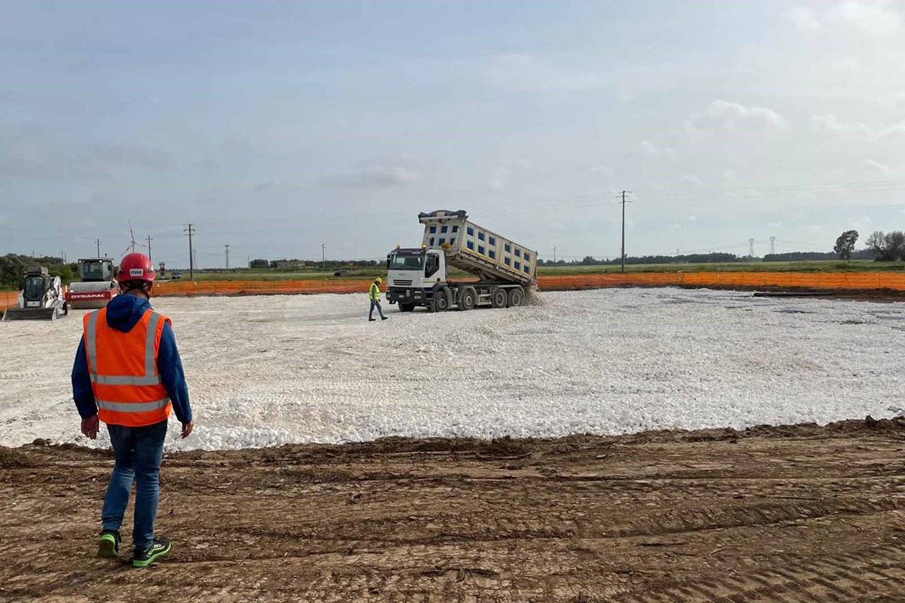 Un cantiere con un camion ribaltabile che scarica materiali su una superficie grigia. I lavoratori supervisionano l'operazione.