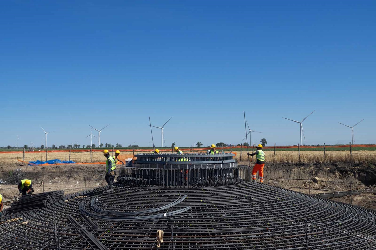 Operai in abbigliamento di sicurezza assemblano una struttura circolare in una giornata di sole, con turbine eoliche visibili sullo sfondo.