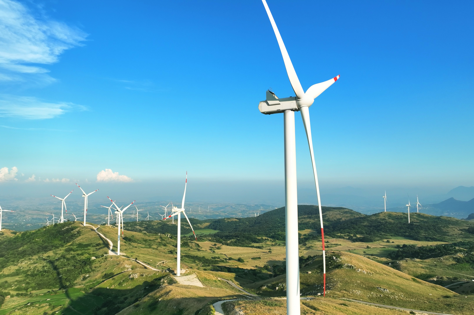 Un paesaggio con molte turbine eoliche su colline verdi sotto un cielo blu chiaro.