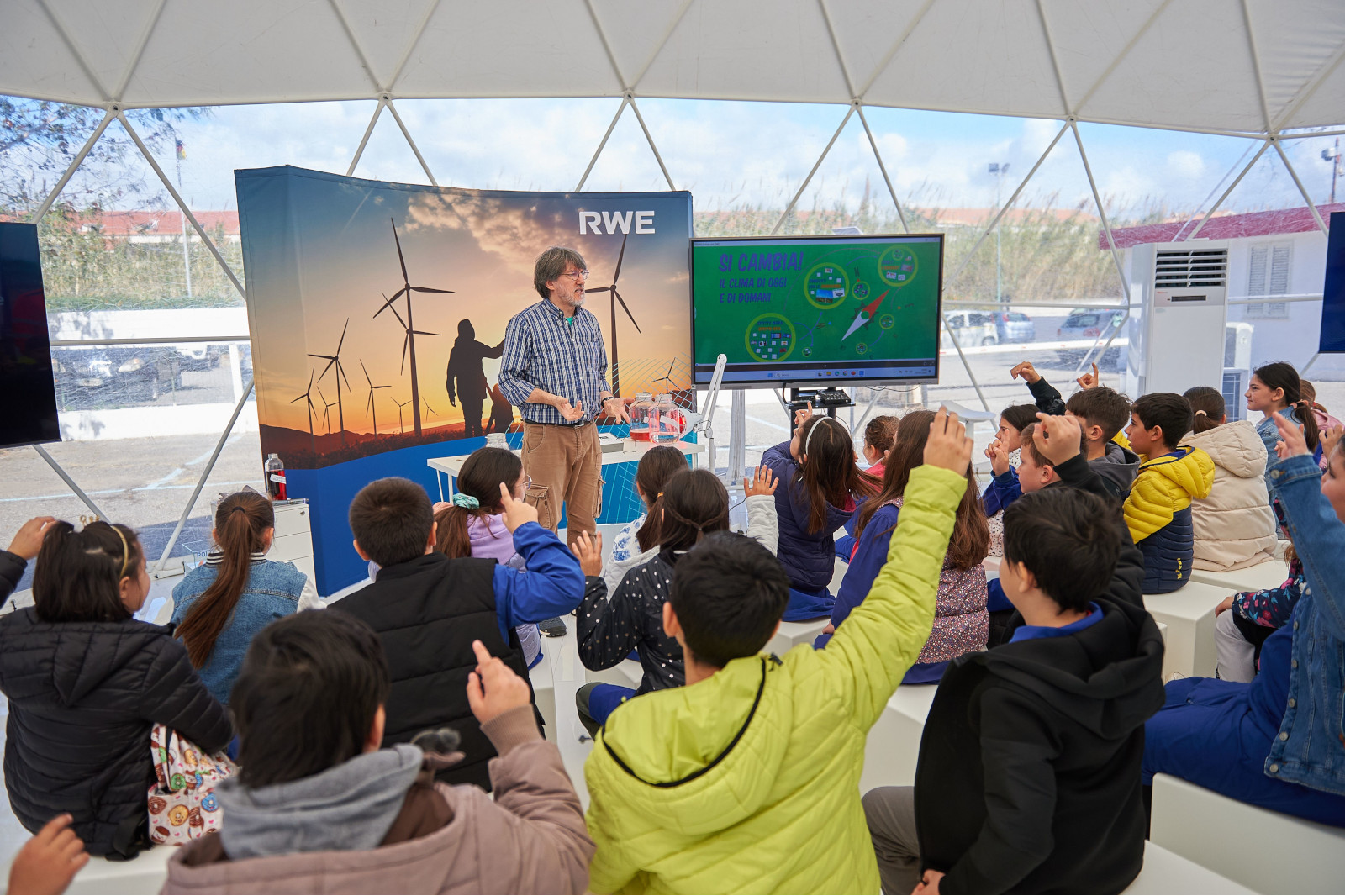 Un insegnante interagisce con i bambini in aula, discutendo di energia rinnovabile con una presentazione digitale e turbine eoliche sullo sfondo.
