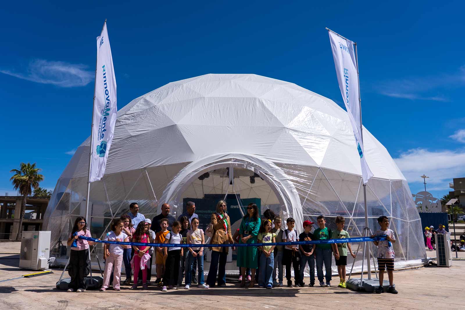 Un gruppo di bambini e adulti si trova davanti a un grande domo geodetico, pronto a tagliare un nastro all'aperto.