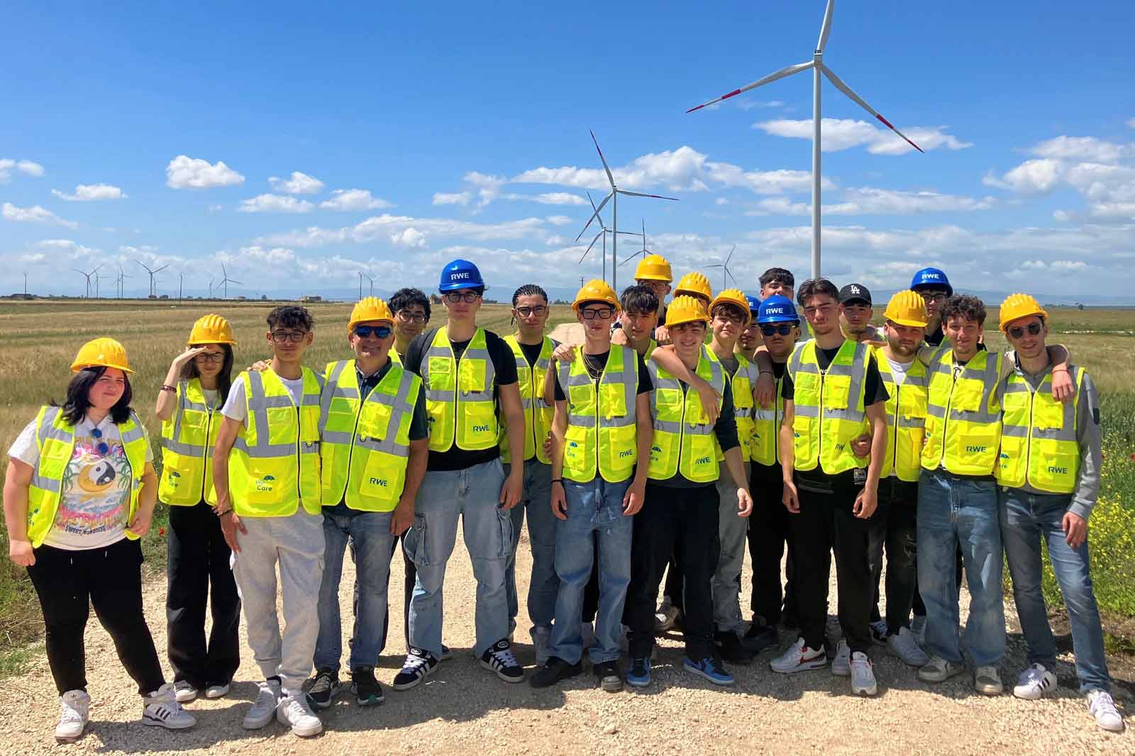 Un gruppo di persone indossa gilet e caschi di sicurezza gialli di fronte a turbine eoliche sotto un cielo azzurro.