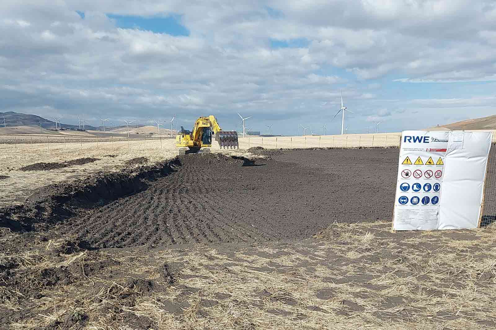 A yellow excavator working on a bare field with wind turbines in the background; a safety notice stands nearby.