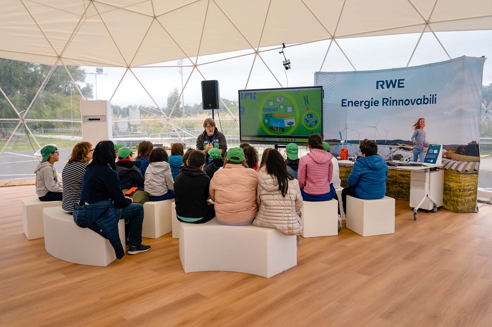 Adulti e bambini su panche bianche dentro una cupola geodetica con pavimento in legno, davanti a uno schermo verde e al cartello "RWE Energie Rinnovabili".
