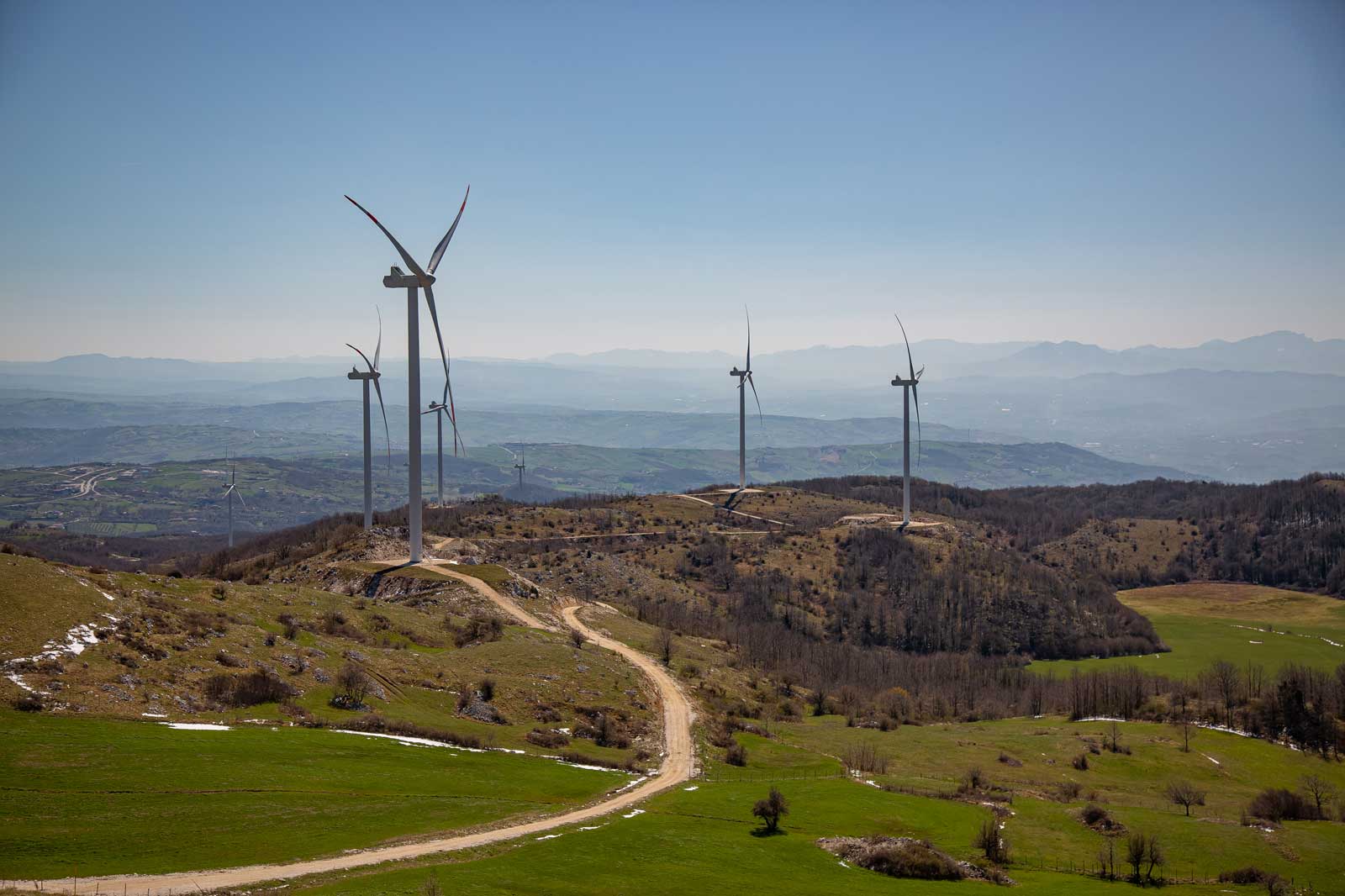 Una vista panoramica di turbine eoliche su una collina verde con cielo blu chiaro e montagne lontane.