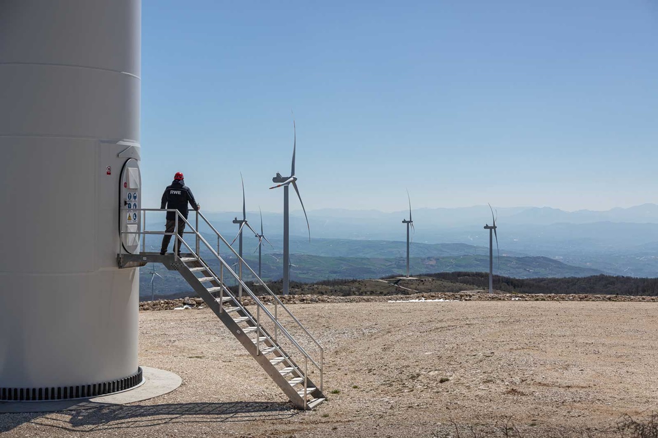 Un tecnico sale una scala metallica accanto a una turbina eolica, con altre turbine sullo sfondo e un cielo sereno.