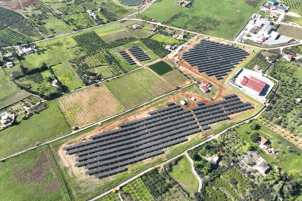 An aerial view of a solar farm with rows of solar panels surrounded by green fields and scattered buildings.