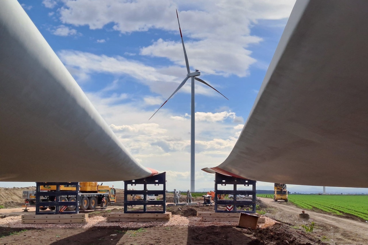 Due pale di turbine eoliche sono posizionate su gru con una turbina eolica sullo sfondo contro un cielo blu con nuvole.