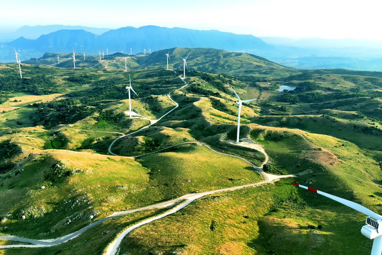 Vista aerea di un paesaggio collinare con turbine eoliche e strade tortuose, circondato da prati verdi.